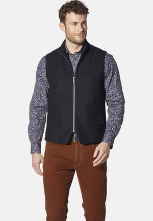 Schwarze ärmellose Weste mit einem Reißverschluss vorne, zwei Fronttaschen und einer glatten Textur, getragen über einem blauen, floral gemusterten Langarmshirt und braunen Hosen.