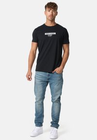 Crna pamučna majica s bijelim logotipom "INDICODE JEANS", uparena s svijetloplavim trapericama i bijelim tenisicama. Ležeran kroj i stil.