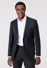 Completo blu scuro con un design aderente, giacca monopetto, camicia bianca e un sottile dettaglio del logo sulla patta. I pantaloni su misura completano il look.
