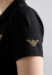 Polo shirt de algodão preto com um design de manga curta, destacado por um detalhe de logotipo com tachas douradas no ombro. Textura suave.