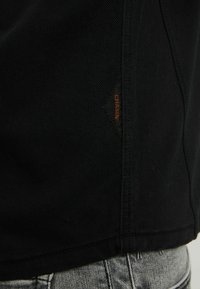 Gros plan sur un tissu noir avec une petite étiquette orange "CHASIN'" cousue près de la couture au-dessus d'un jean denim gris clair.
