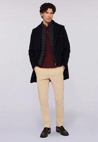 Černý kabát přes tmavou péřovou bundu, vínové polo, béžové kalhoty a tmavé boty. Kabát má na míru šitý střih a hladkou texturu.