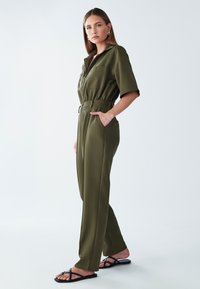 Mono verde oliva con mangas cortas, cierre frontal con cremallera, cintura elástica y bolsillos laterales. Combinado con sandalias negras con tiras. Textura suave.