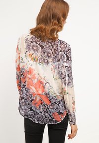 Blouse florale transparente présentant un mélange de couleurs sombres et vives, des motifs floraux complexes et une coupe ample. Manches longues et ourlet arrondi.