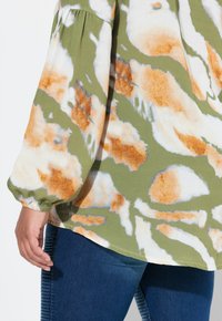 Blusa verde con motivo astratto arancione e bianco, maniche lunghe a sbuffo e polsini elasticizzati. Abbinata a jeans denim blu scuro.