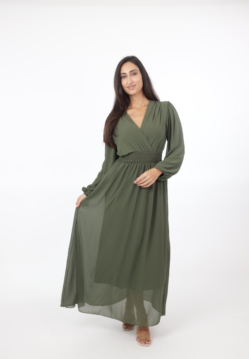 Vrouw met lang donker haar die een lange maxi-jurk met lange mouwen in olijfgroen draagt, houdt de rok licht vast terwijl ze staat tegen een witte achtergrond.