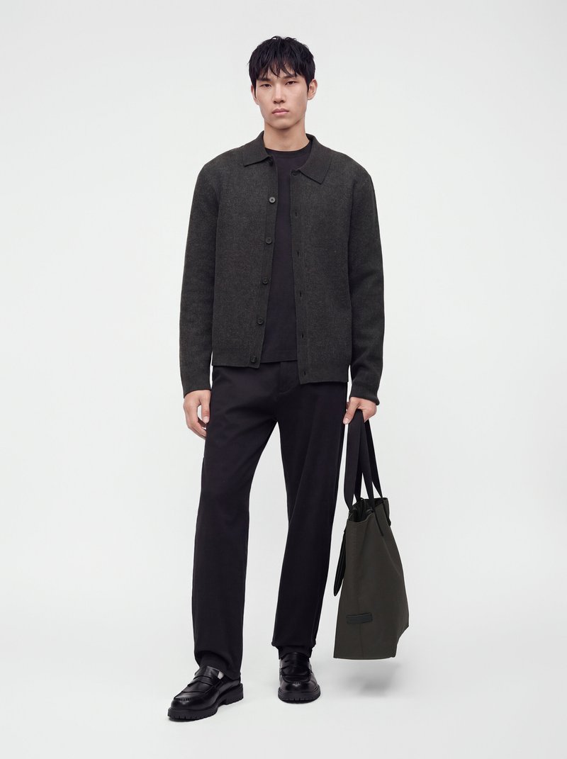 Un homme porte une veste anthracite boutonnée sur le devant, un pantalon et des chaussures noirs. Il tient un sac cabas sombre devant un fond de studio blanc uni.
