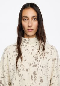 Maglioncino beige a maglia con colletto alto, tessuto testurizzato, caratterizzato da macchie scure e un design semplice. Nessun hardware o accenti visibili.