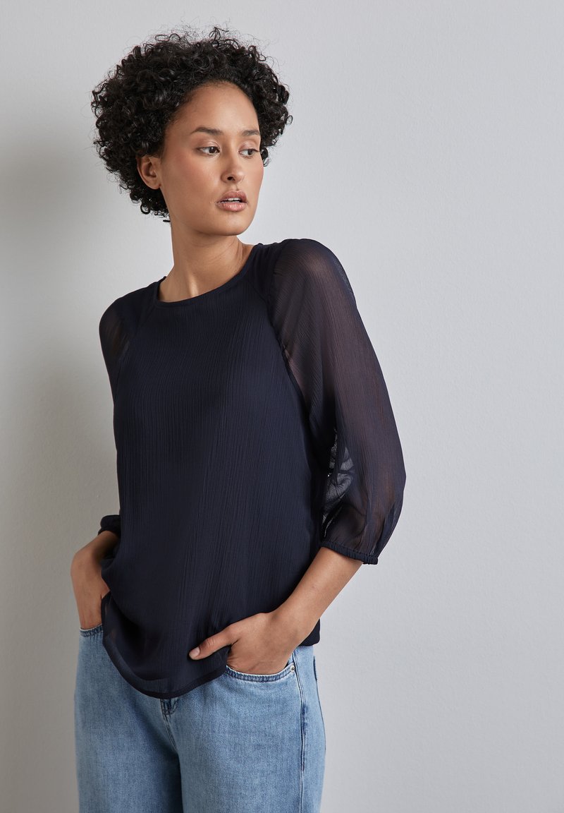 Blusa blu navy con texture plissettata, maniche a palloncino trasparenti e scollo rotondo. Abbinata a jeans azzurri, mani nelle tasche.
