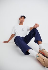 White t-shirt with graphic print, navy blue trousers, silver cap, and beige sneakers with colorful accents. Plain background.