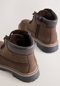 Braune Leder-Stiefeletten mit weichem Innenfutter, seitlichem Reißverschluss, strukturierter Oberfläche, braunen Akzenten und einer robusten schwarzen Gummisohle.