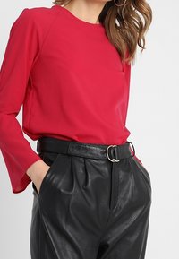 Frau trägt eine rote Bluse mit langen Ärmeln und Glockenärmel, die in schwarze Lederhosen mit hoher Taille und einem Doppelschnallen-Gürtel gesteckt ist, Hände in den Taschen.