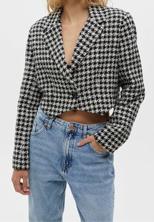 Vrouw draagt een zwart-witte houndstooth cropped blazer met één zwarte knoop, gecombineerd met lichtblauwe high-waisted jeans.