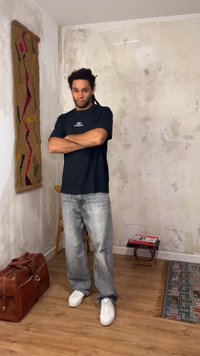 Άντρας με σταυρωμένα χέρια, που φοράει μαύρο μπλουζάκι και ξεθωριασμένο τζιν, στέκεται σε ένα δωμάτιο με παλιά υφή στον τοίχο και ξύλινο πάτωμα.
