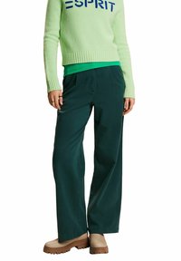 Zelený svetr s modrým nápisem, spárovaný s tmavě zelenými širokými kalhotami a béžovými botami. Tkaniny vypadají pleteně a hladce. Jednoduchý design.