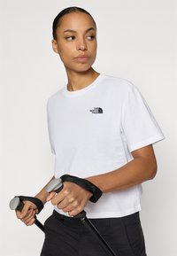 Λευκό βαμβακερό T-shirt με κοντά μανίκια, με λογότυπο στο αριστερό στήθος σε μαύρο χρώμα. Το μοντέλο κρατάει μπαστούνια πεζοπορίας με γκρι λαβές.