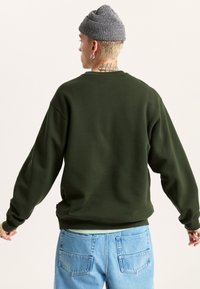 Sudadera verde oscura con hombros caídos, cuello redondo y puños de canalé, combinada con jeans azul claro que tienen dos bolsillos traseros.
