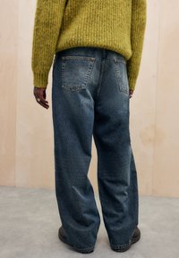 Jeans azuis largos e desbotados com uma cintura alta e bolsos traseiros, combinados com um suéter texturizado e canelado na cor mostarda.