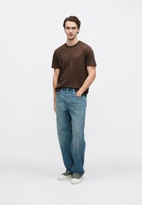 Καφέ βαμβακερό μπλουζάκι με μικρό πορτοκαλί λογότυπο, συνδυασμένο με ανοιχτό μπλε τζιν παντελόνι. Το μοντέλο φοράει πράσινα αθλητικά παπούτσια. Απλός, καθημερινός σχεδιασμός.