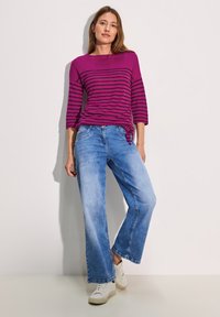 Violetti ja musta raidallinen pusero, jossa on 3/4-hihat, yhdistettynä vaaleansinisiin leveälahkeisiin farkkuihin ja valkoisiin tennareihin. Suorat pinnat ja rento muotoilu.