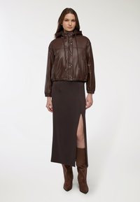 Giubbotto in pelle marrone cropped con cappuccio, chiuso con bottoni, abbinato a una lunga gonna marrone con uno spacco laterale e stivali marroni al ginocchio.