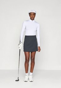 Λευκό μακρυμάνικο τοπ με ψηλό λαιμό, γκρι πλισέ φούστα, λευκό καπέλο, μπαστούνι του γκολφ και λευκά αθλητικά παπούτσια σε ουδέτερο φόντο.