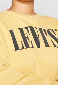 Sudadera amarilla de tejido suave, con un gran logo negro de "LEVI'S". Cuello acanalado, corte relajado y mangas ligeramente gatheradas.