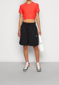 Roter Cropped-Top mit Nike-Logo, schwarze Shorts mit Kordelzug, weiße Umhängetasche und graue High-Top-Sneaker mit roten Akzenten.