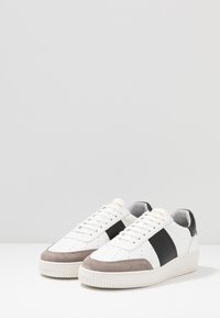 Vita sneakers med en slät läderöverdel med ett svart rand, grå mocka tå och texturerad gummisula. Snördesign med ventilationshål.