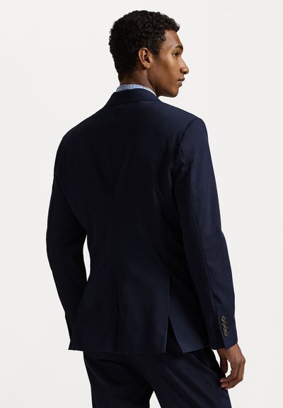 Blazer azul escuro feito de tecido suave, com corte ajustado, lapelas picadas e punhos com três botões. Abertura nas costas para facilitar o movimento.