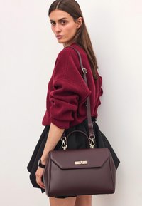 Bordowa skórzana torebka o strukturalnym designie, ze złotymi akcentami, górnym uchwytem i odpinanym paskiem na ramię, w zestawieniu z wygodnym swetrem.