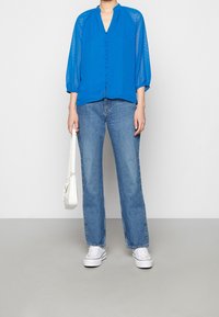 Blå, halvgenomskinlig blus med trekvartsballongsärmar och en nyckelhålsknäppning. Kombinerad med ljusblå raka jeans och vita sneakers.