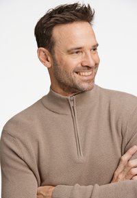 Jersey de punto color beige con un patrón texturizado y un cuello alto. Presenta una cremallera en la parte frontal y puños de canalé.