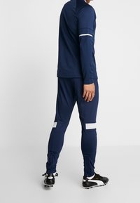 Marineblauwe atletische trainingsjurk met lange mouwen en taps toelopende broek; met witte strepen op de mouwen en zijkanten van de broek; gemaakt van glad, rekbaar materiaal.