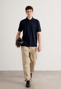 Camisa polo azul marinho, de manga curta, com logo no peito; calças bege, de corte descontraído; sapatilhas pretas; segurando uma bolsa preta; fundo neutro.