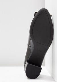 Svart lackläder skon med spetsig tå och klack med block, som visar varumärket "högl" präglat på undersidan.