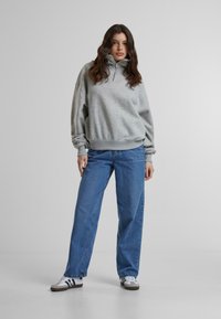 Sudadera gris con cremallera y cuello alto, combinada con jeans anchos de color azul y zapatillas blancas con detalles oscuros. Tela suave y casual.