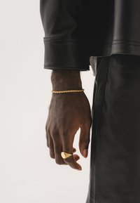 Veste en cuir noir avec une finition texturée ; main portant un bracelet en or torsadé et une bague en or ornée.