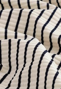 Teksturoidu kangasta, jossa on kerma- ja tummansinisiä raitoja, kuisti-kuviointi ja vaihteleva leveys, luoden kerroksellisen, kudotun ilmeen.