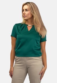 Szmaragdowozielona bluzka z krótkim rękawem i dekoltem w kształcie litery V, z akcentem w postaci złotego łańcuszka przy kołnierzyku oraz gładką, błyszczącą teksturą.