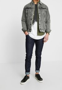 Džínová bunda, šedá s vybledlými detaily; přes ní vrstvené tričko s potiskem ve tmavě zelené a bílé; tmavé džíny s ohrnutými manžetami; černé tenisky.