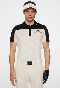 Herrarnas golfskjorta i beige med svarta accenter. Har ett hexagonmönster, krage och logotyp. Bärs med krämfärgade byxor och en svart handskar.