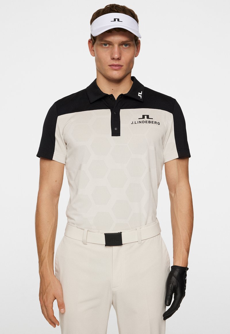 Herrarnas golfskjorta i beige med svarta accenter. Har ett hexagonmönster, krage och logotyp. Bärs med krämfärgade byxor och en svart handskar.