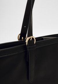 Bolso de mano de imitación cuero negro con dos asas superiores, acentos de hardware circular dorado y detalles de costura. Cerrado, sin mostrar características interiores.