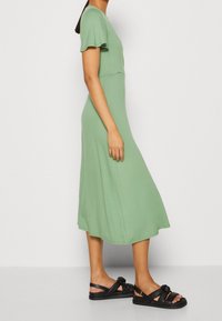 Vihreä midi-mekko lyhyillä, liehuvilla hihoilla, valmistettu pehmeästä kankaasta. Yhdistetty mustiin litteisiin sandaaleihin, joissa on solkityyppinen yksityiskohta ja paksut hihnat.