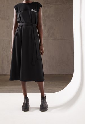 Abito midi nero con top aderente, design senza maniche e cintura in vita. Il materiale è liscio con una finitura opaca. Indossato con stivaletti neri alla caviglia.