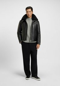 Schwarze Lederjacke mit großem Lammfellkragen, grauer Strickpullover, schwarze Hose und schwarze Sneaker mit weißen Sohlen.