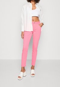 Pantalon en coton rose à coupe ajustée, associé à un débardeur court blanc et une chemise blanche ouverte à boutons ; sandales à plateforme blanches.