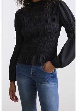 Frau trägt eine schwarze, geraffte Langarmbluse mit Rüschensaum und blaue Jeans, eine Hand in der Tasche, steht vor einem einfarbigen Hintergrund.