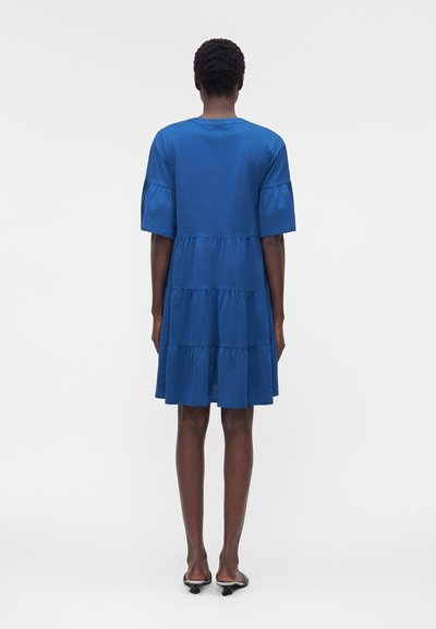 Γυναίκα όρθια, φοράει ένα φαρδύ μπλε φόρεμα με βαθμίδες και μανίκια μέχρι τον αγκώνα και μαύρα πέδιλα με τακούνι και λουράκι πίσω, με την πλάτη στραμμένη.